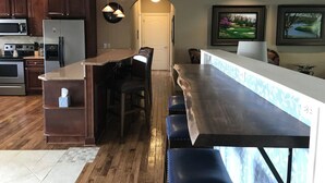 Une cuisine moderne avec des meubles en bois foncé, des appareils électroménagers en acier inoxydable, un îlot central doté de tabourets de bar et une vue sur l’espace de vie à travers une arche.