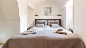 A neatly made bed with a headboard, a bedside table with a lamp, and a wall-mounted shelf with various items. - Villa Cucunci by BarbarHouse (Ostuni)