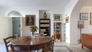 A dining room with a wooden table, chairs, a vase with flowers, a bookshelf, a clock, and framed pictures on the walls. - 2947 Aia del Convento by BarbarHouse (Marittima)