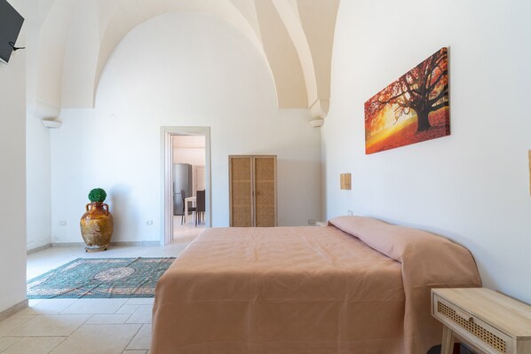 Un dormitorio con cama, un jarrón grande, un cuadro y una puerta que da a otra habitación.