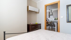 A bedroom with a bed, a dresser, and a small desk with a chair. There is a kitchenette visible through an open door.