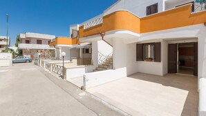 A residential building with a white facade and orange accents, featuring a staircase leading to a balcony, a visible kitchen area, and a parked car in the background.
