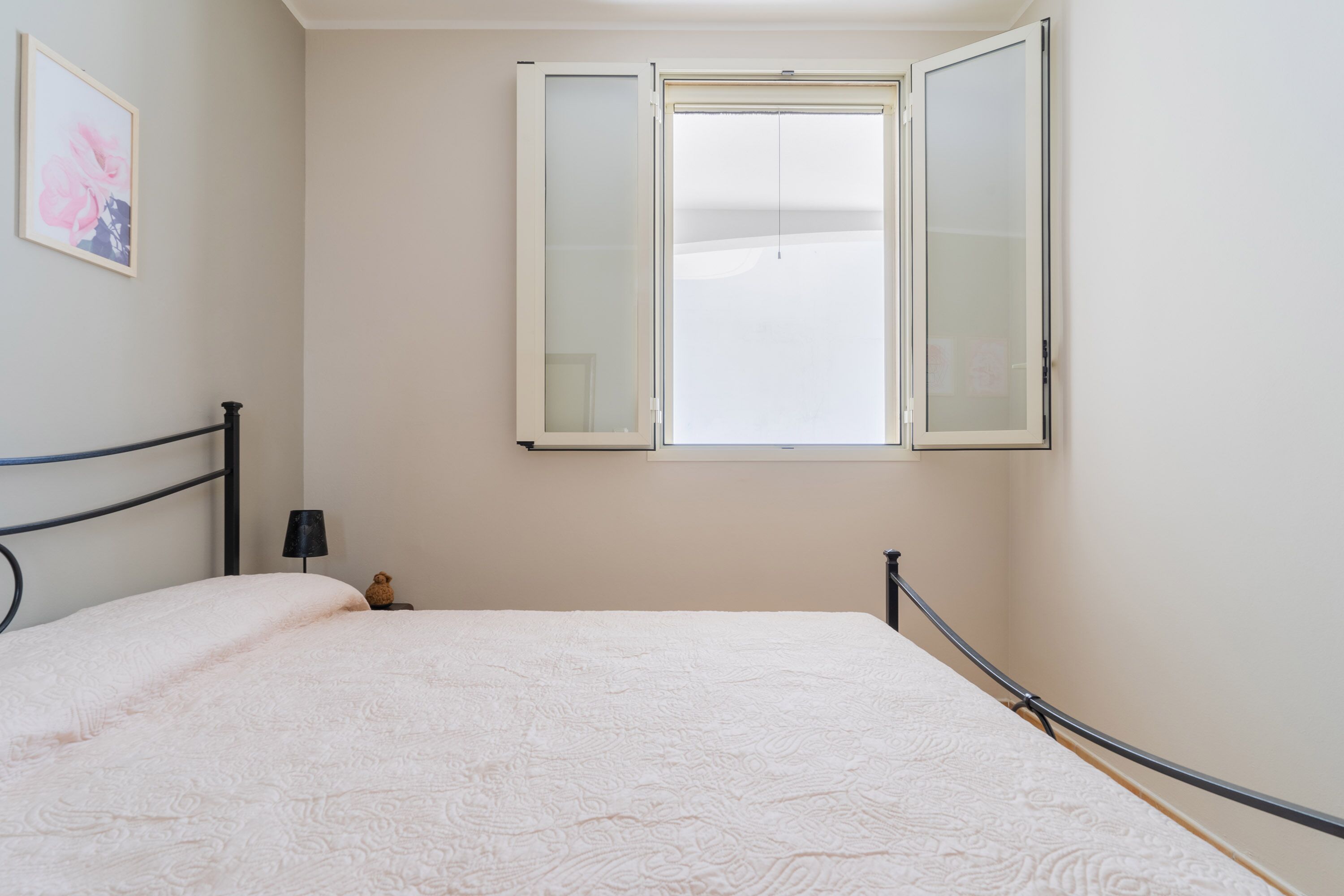 Une chambre avec un lit simple, une fenêtre dont les volets sont ouverts, une petite table de chevet avec une lampe et un tableau encadré accroché au mur.