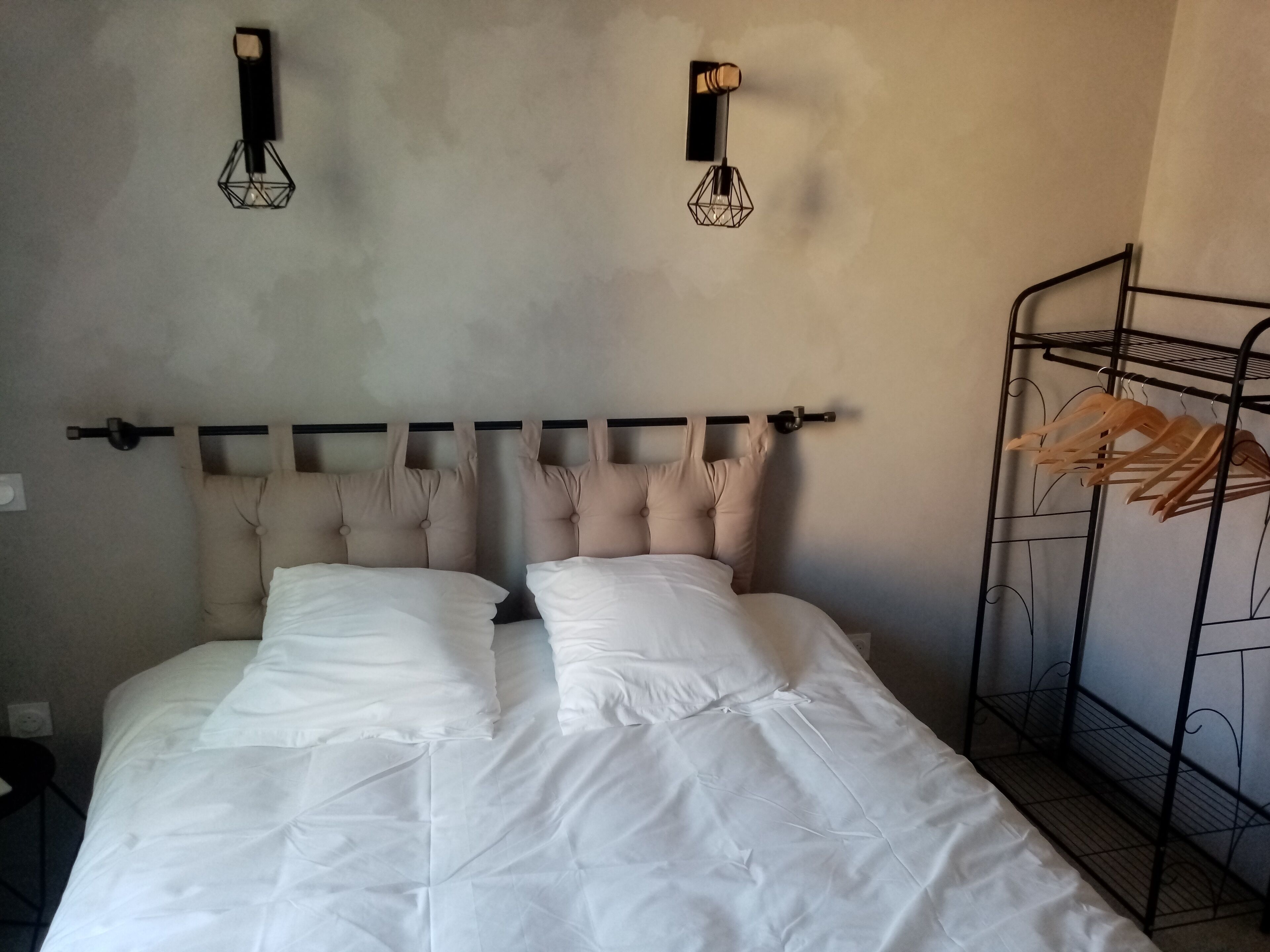 A bed with a headboard, two wall-mounted light fixtures, and a metal shelf unit with clothes.