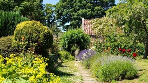 Un sendero de jardín que conduce a un edificio de piedra con techo de tejas, rodeado de plantas y follaje florido.