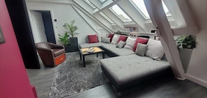 A modern loft-style living room with a grey sectional sofa, a brown leather armchair, and a wooden coffee table. There is a skylight above the sofa and a large potted plant to the left.