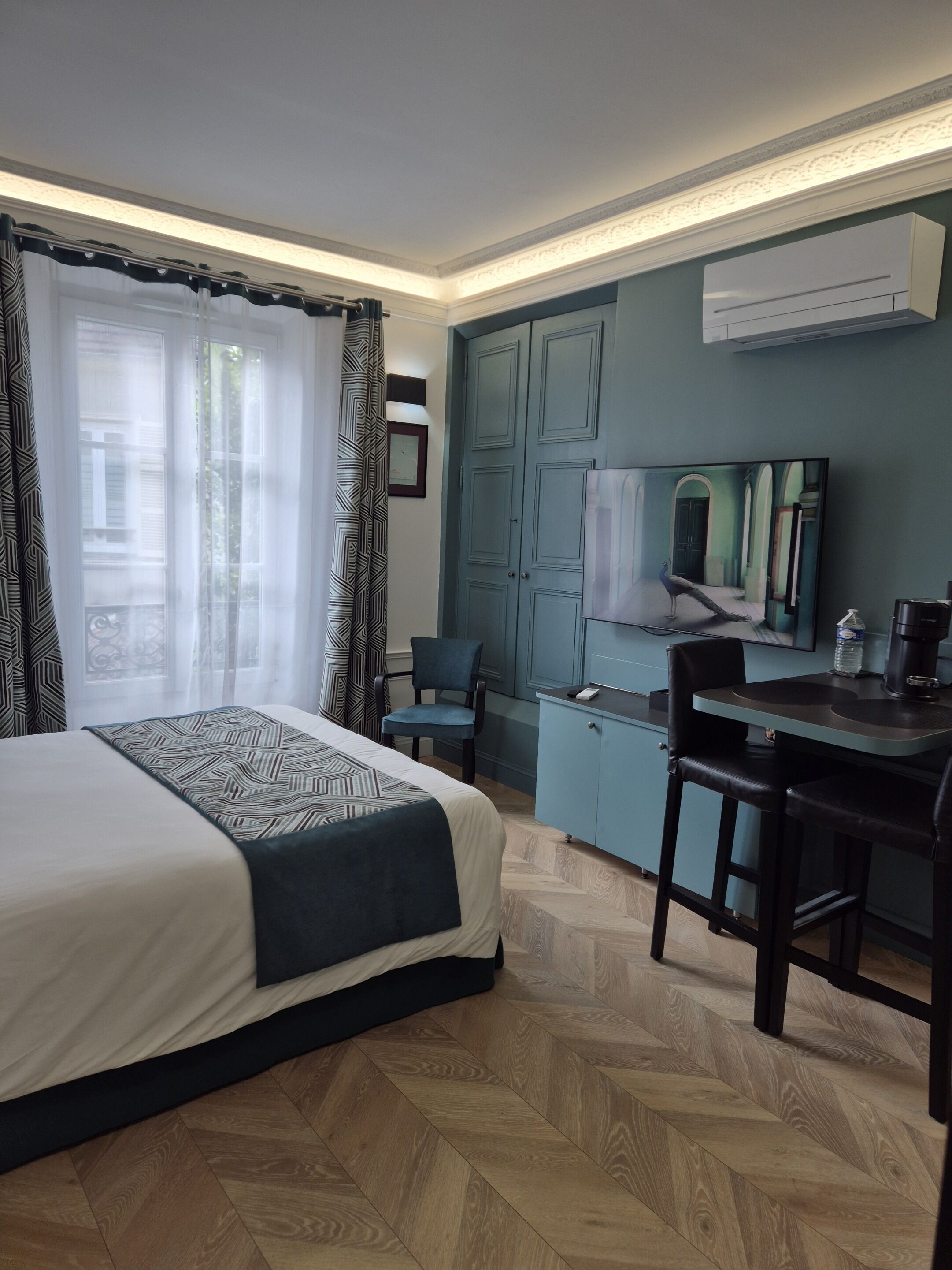 Une chambre d’hôtel moderne avec un grand lit, un bureau et une chaise. On y trouve une fenêtre avec des rideaux, un miroir et un appareil de climatisation fixé au mur.