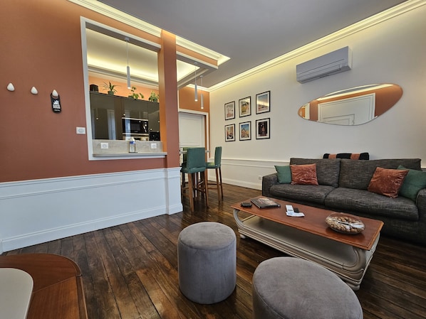A modern living room with a grey sofa, ottomans, a wooden coffee table, and a kitchen area visible through a large window.