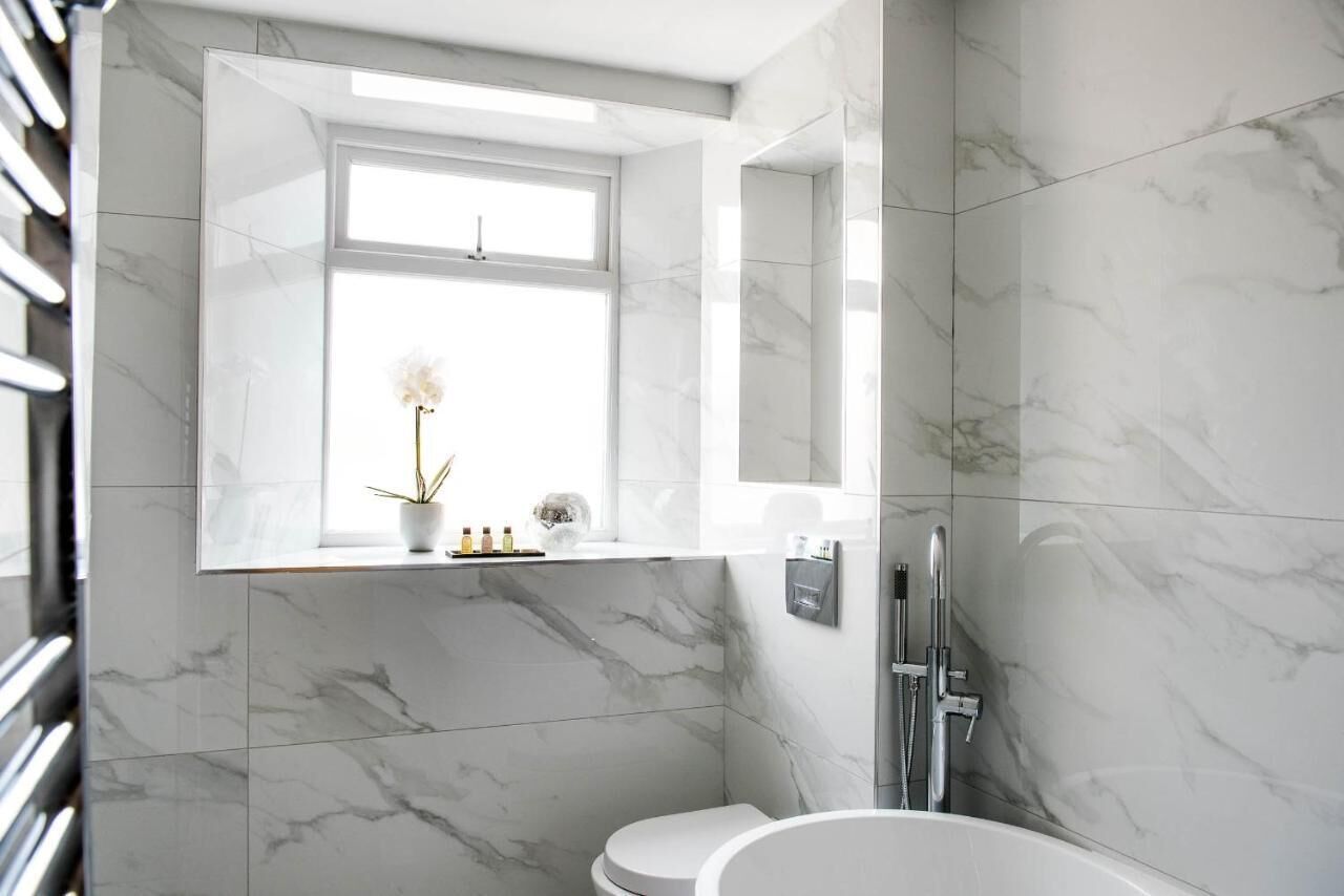Un baño moderno con suelo de mármol, inodoro blanco, bañera independiente y una ventana con una orquídea en maceta.