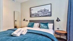 Et hotelværelse med en stor seng, sengegavl, et natbord, en lampe, et billede i ramme på væggen og et vindue med gardiner.