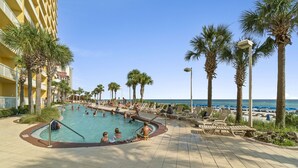 Zona de piscina con sillas de descanso y palmeras, junto a una playa y un edificio de hotel.