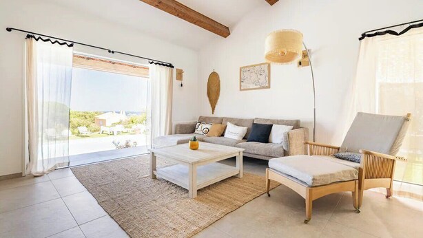 Ein geräumiges Wohnzimmer mit Sofa, Couchtisch und Sessel. Ein großes Fenster mit Vorhängen, eine Wandlampe und ein gerahmtes Kunstwerk sind ebenfalls vorhanden.