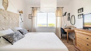 Ein Schlafzimmer mit einem großen Bett, einem Schreibtisch, einem Stuhl, einem Fernseher und Blick ins Freie durch das Fenster.