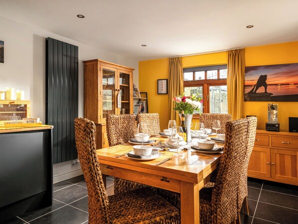 A dining area with a wooden table set for four, surrounded by woven chairs. A cabinet with glass doors is to the left, and a window with curtains is to the right.