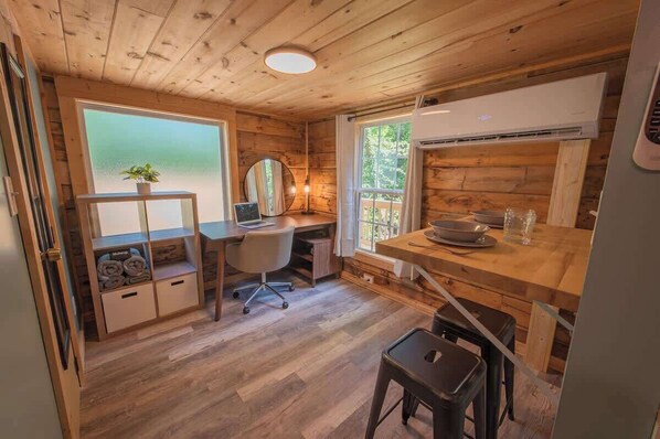 Ein gemütlicher Raum mit einem Holztisch, einem Stuhl, einem kleinen Hocker, einem Spiegel und einem Fenster mit Blick ins Grüne.
