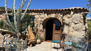 A stone building with a tiled roof, a wooden door, and a patio area with chairs and cacti. - Domaine de Piscia (Figari)