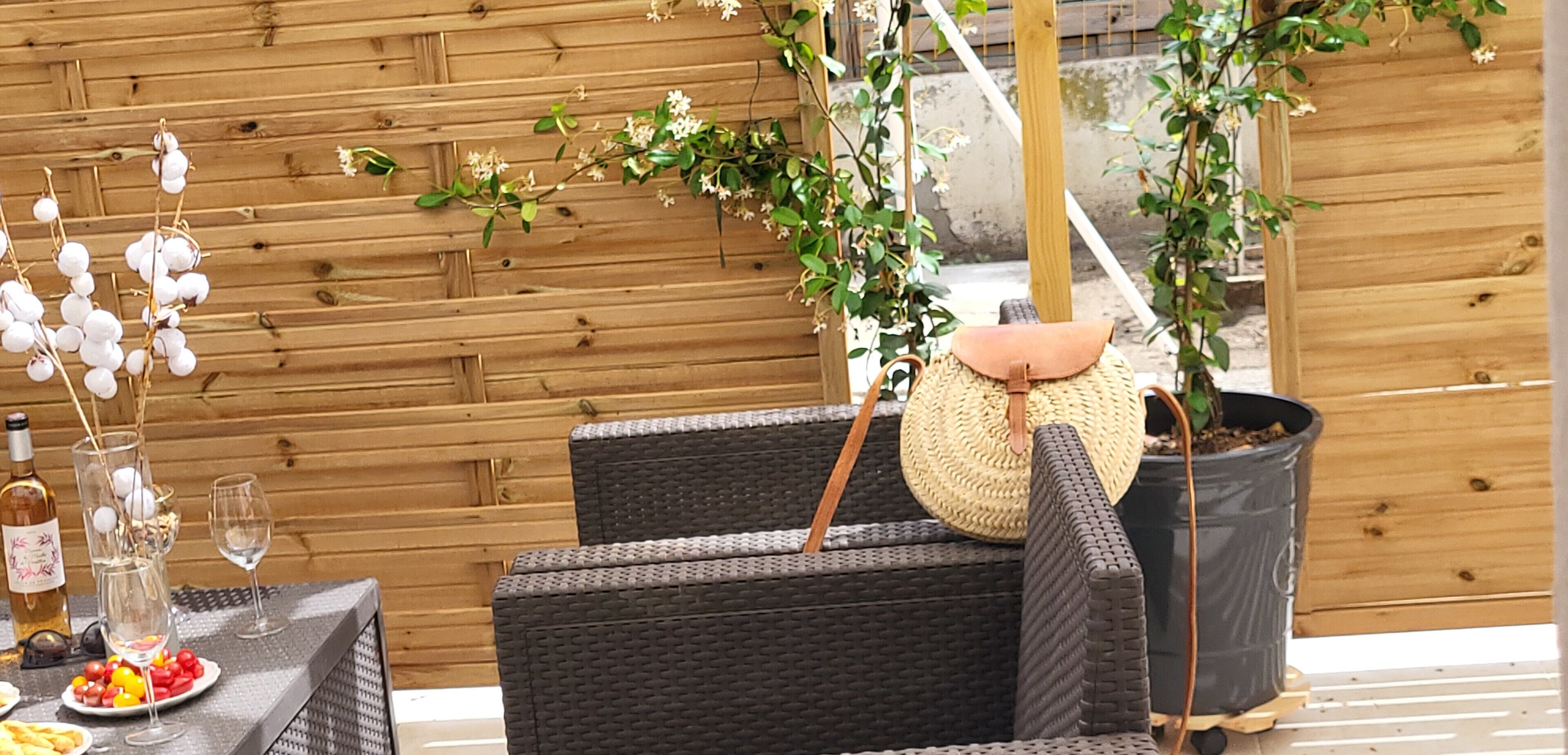 Un coin salon extérieur aménagé avec des meubles en osier, une table dressée pour le repas, une plante en pot et une bouteille de vin.
