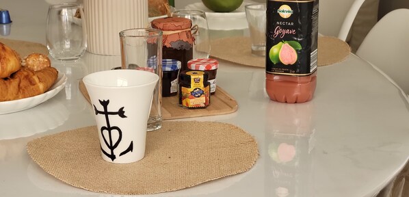 A table with a bottle of Goyave nectar, a jar of jam, a jar of honey, a croissant, and a mug with a symbol.
