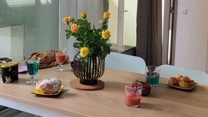 Comedor con mesa dispuesta para dos personas, con un jarrón de flores amarillas, un vaso de bebida rosa y diversos pasteles.