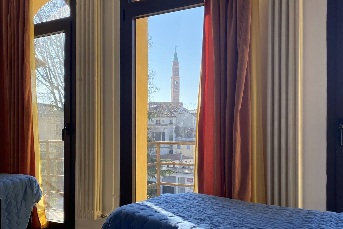 Een hotelkamer met twee bedden, grote ramen met gordijnen en uitzicht op een gebouw met een toren.