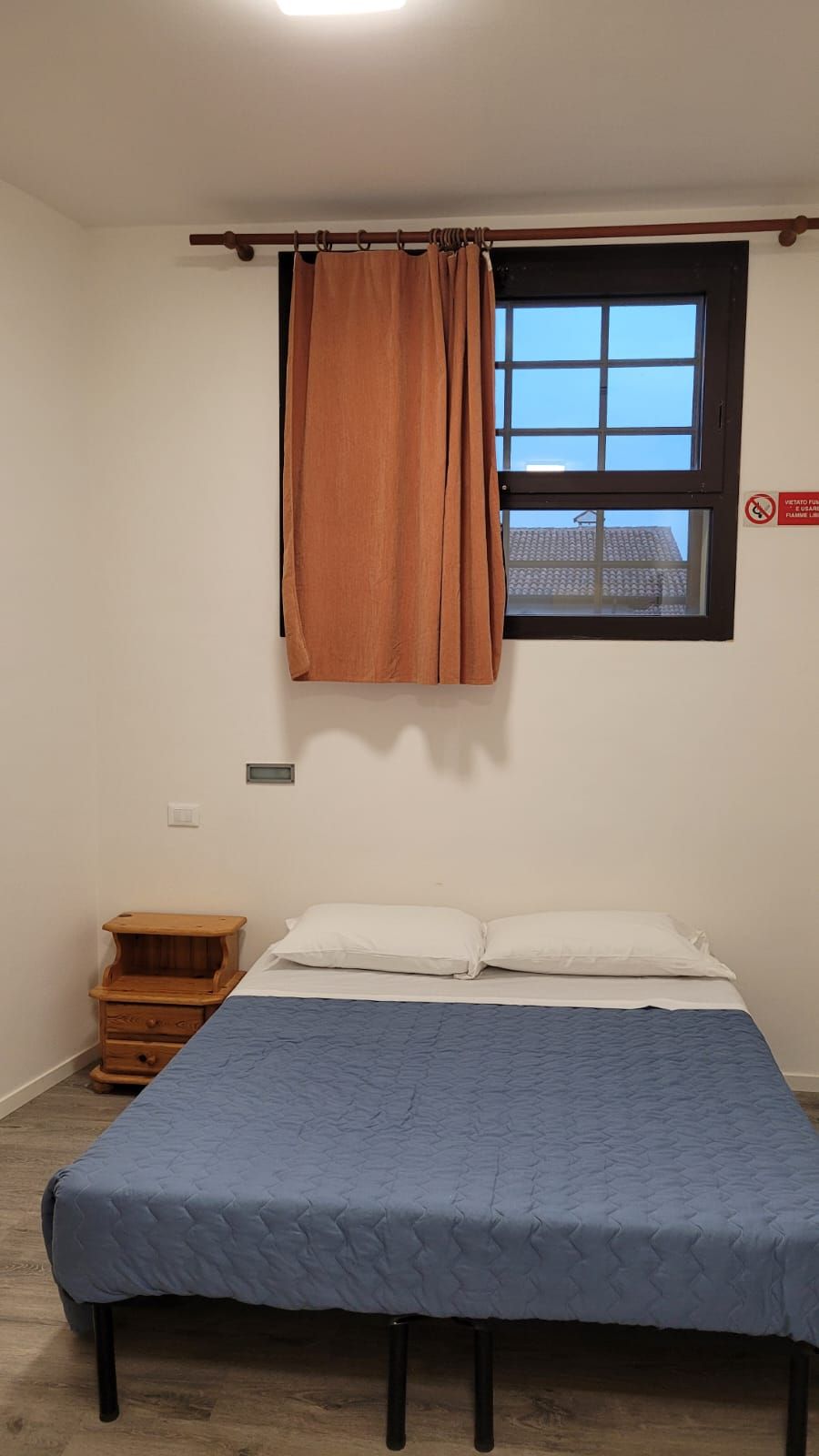 Une chambre à coucher avec un lit, une fenêtre avec un rideau, une petite table de chevet en bois et un luminaire fixé au mur.