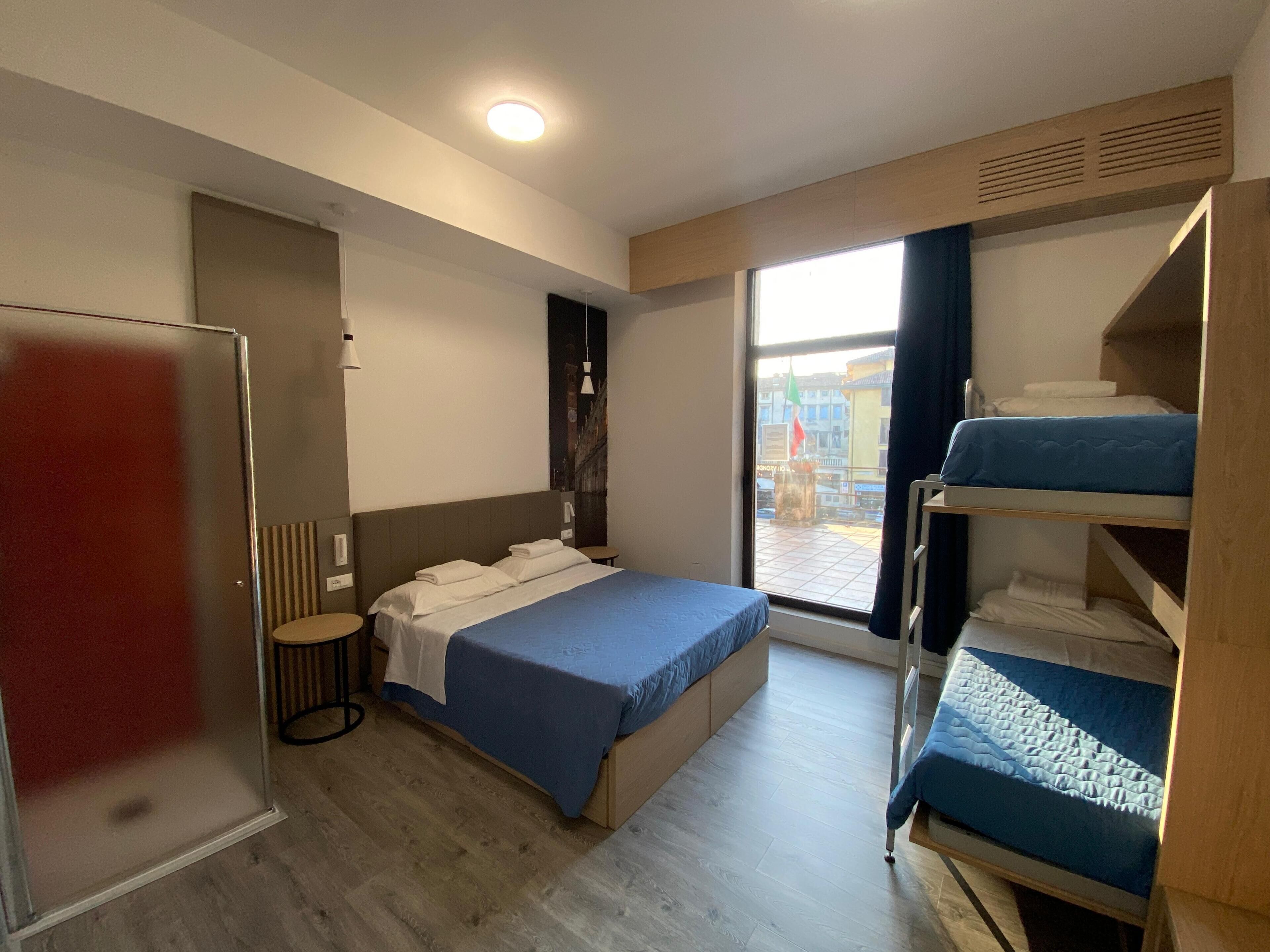 A hotel room with a bed, a small table, a wardrobe, and a window with a view of the outside.