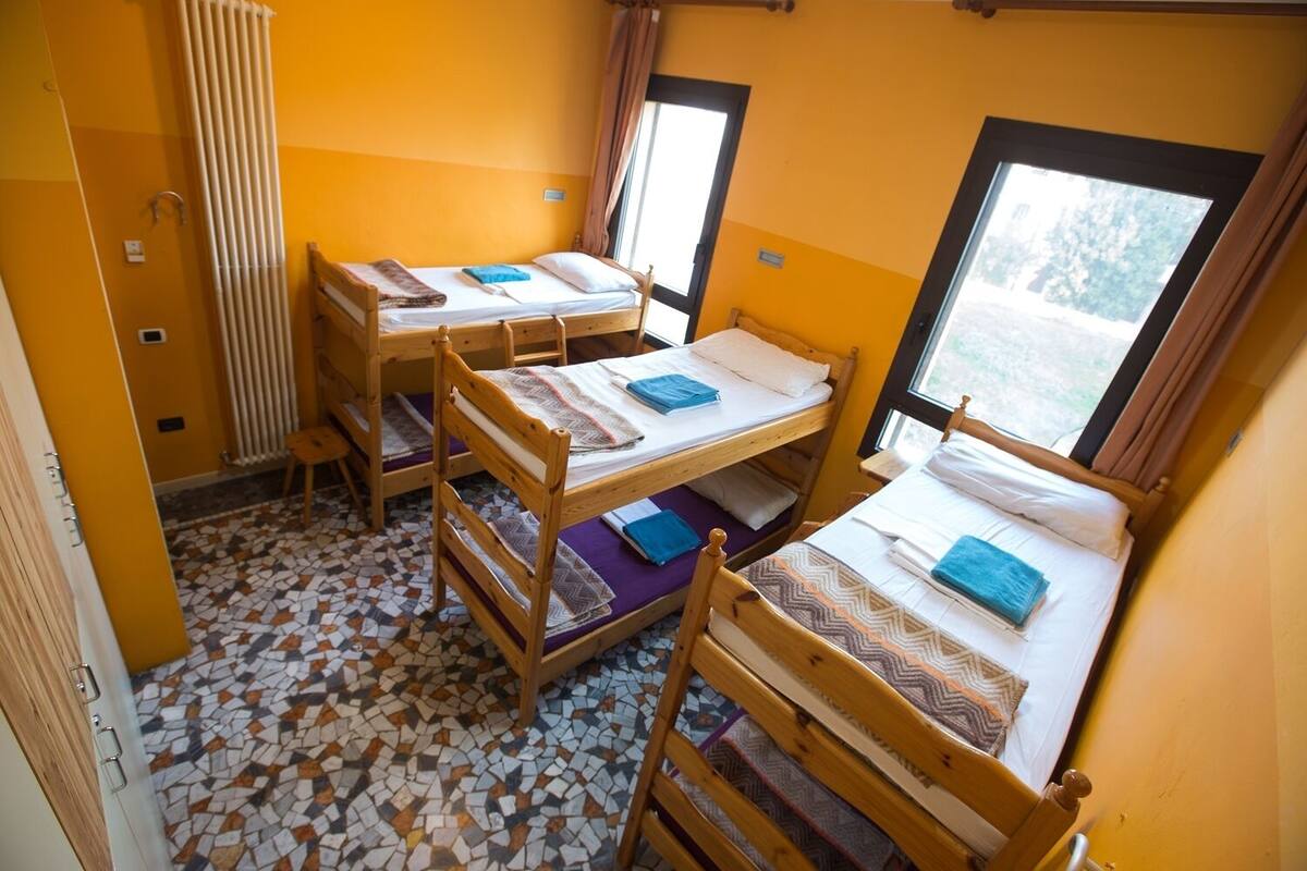 Een slaapzaal met vier stapelbedden, een radiator, een raam met gordijnen en een betegelde vloer.