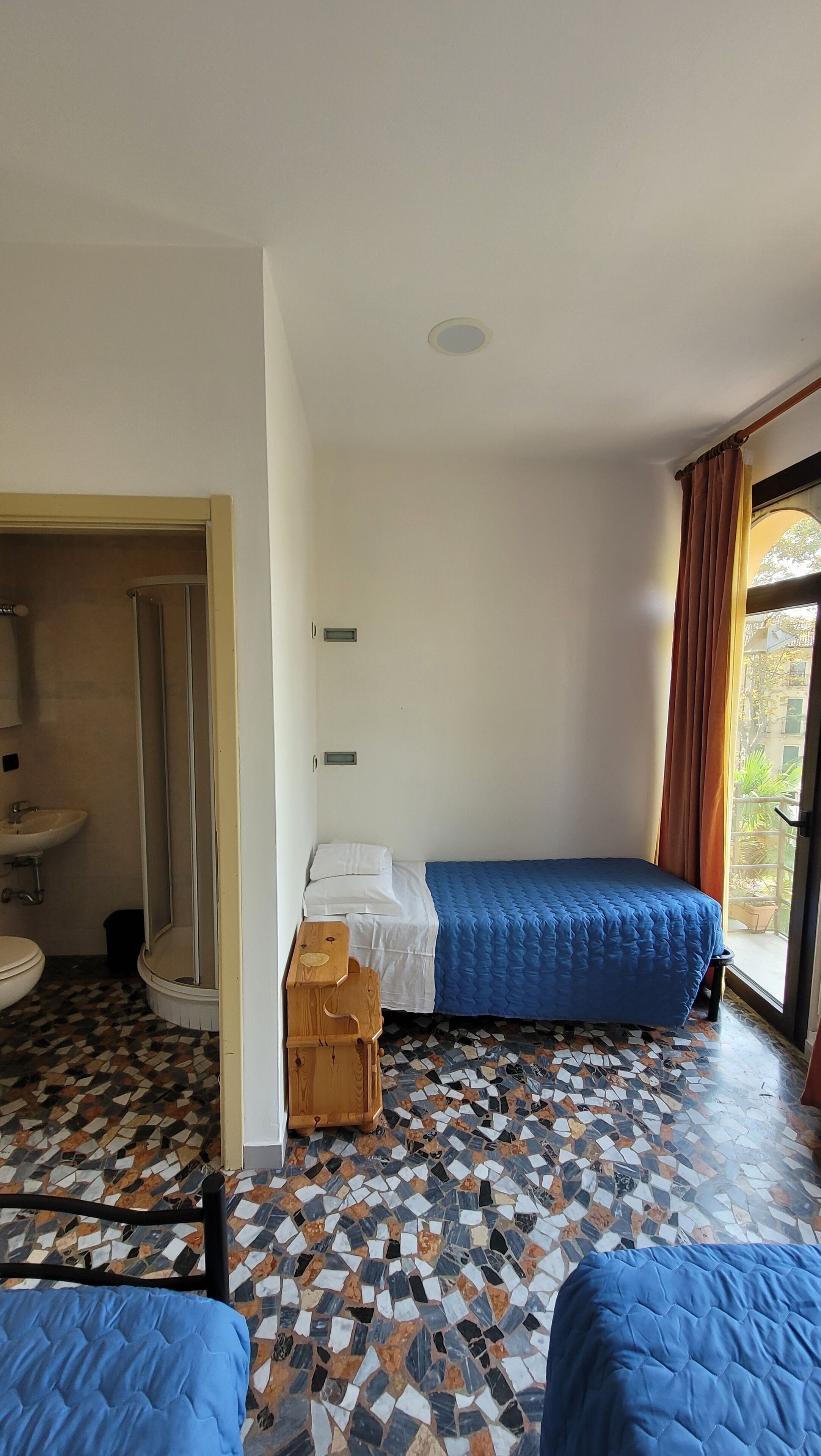 A room with a bed, a small wooden table, a sink, and a shower.