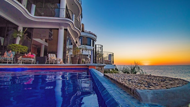 Vue sur une piscine d’hôtel, avec le soleil couchant sur l’océan, des espaces de détente en plein air et une vue sur la mer.