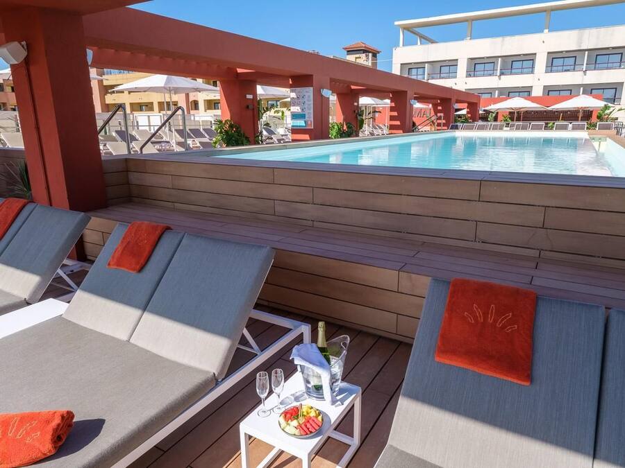 Zona junto a la piscina con sillones, una mesa con bebidas y aperitivos, y vistas a la piscina y los edificios del hotel.