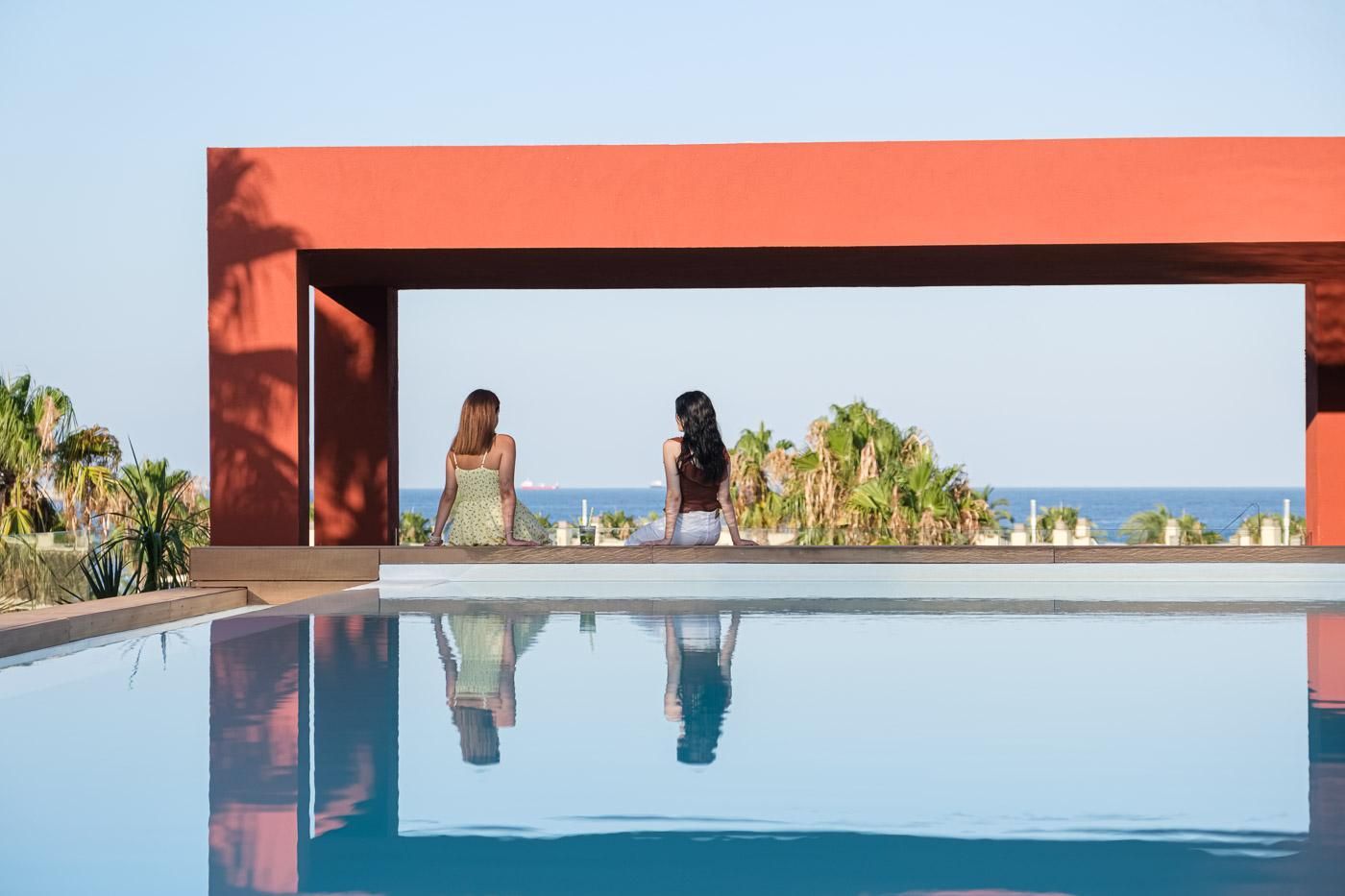 Two women sitting by a pool with a view of the ocean and palm trees.