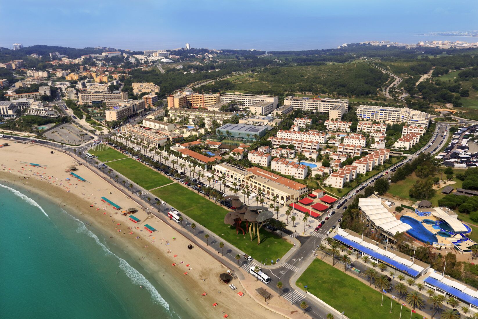 Vista aérea de un complejo turístico frente al mar, con playa, edificios de hotel y una piscina.