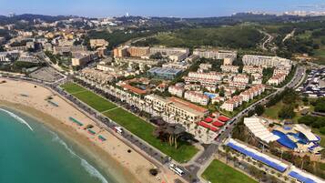 Vista aérea de un complejo turístico frente al mar, con playa, edificios de hotel y una piscina.