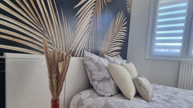 Ein Schlafzimmer mit einem Bett, einem Nachttisch, einer Vase mit Trockenpflanzen und einer Wand mit Blätermuster.