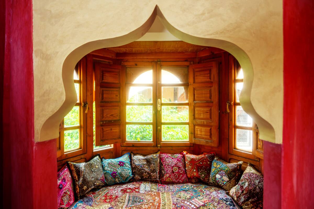 A cozy room with a wooden bench and colorful cushions, large windows, and a unique arched ceiling.