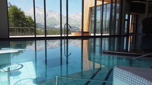 A modern indoor swimming pool with clear glass walls, a view of mountains, and a wooden deck area. - Superdevoluy545S (Le Dévoluy)