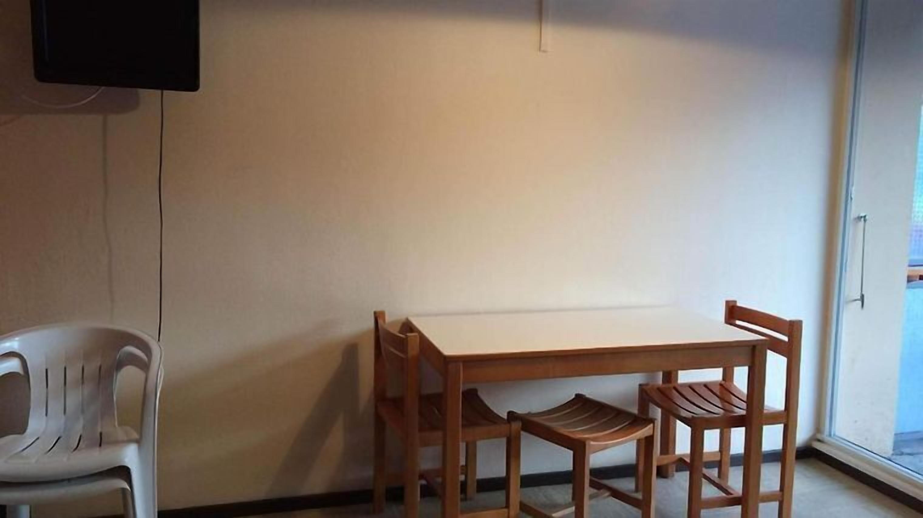 Quarto com uma mesa de madeira e duas cadeiras, uma cadeira de plástico branca e uma TV fixada na parede.