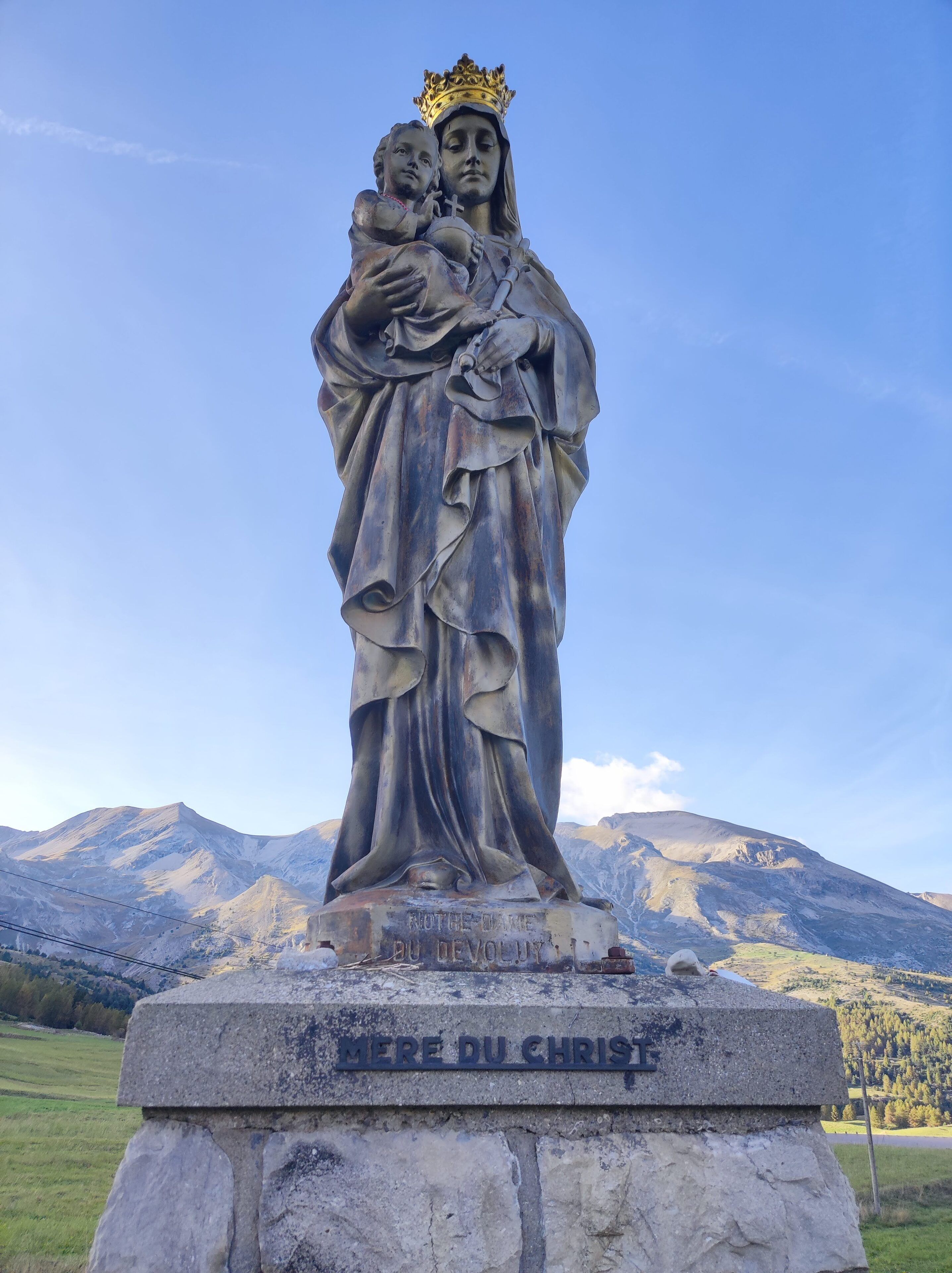 A statue of the Virgin Mary with a child, titled "MERE DU CHRIST," set against a mountainous backdrop.