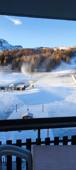 A view from inside a building looking out at a snowy landscape with a ski lift, a wooden building, and a forested mountain in the background.