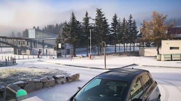 A snow-covered parking area with a modern building, a pedestrian bridge, and a row of trees.