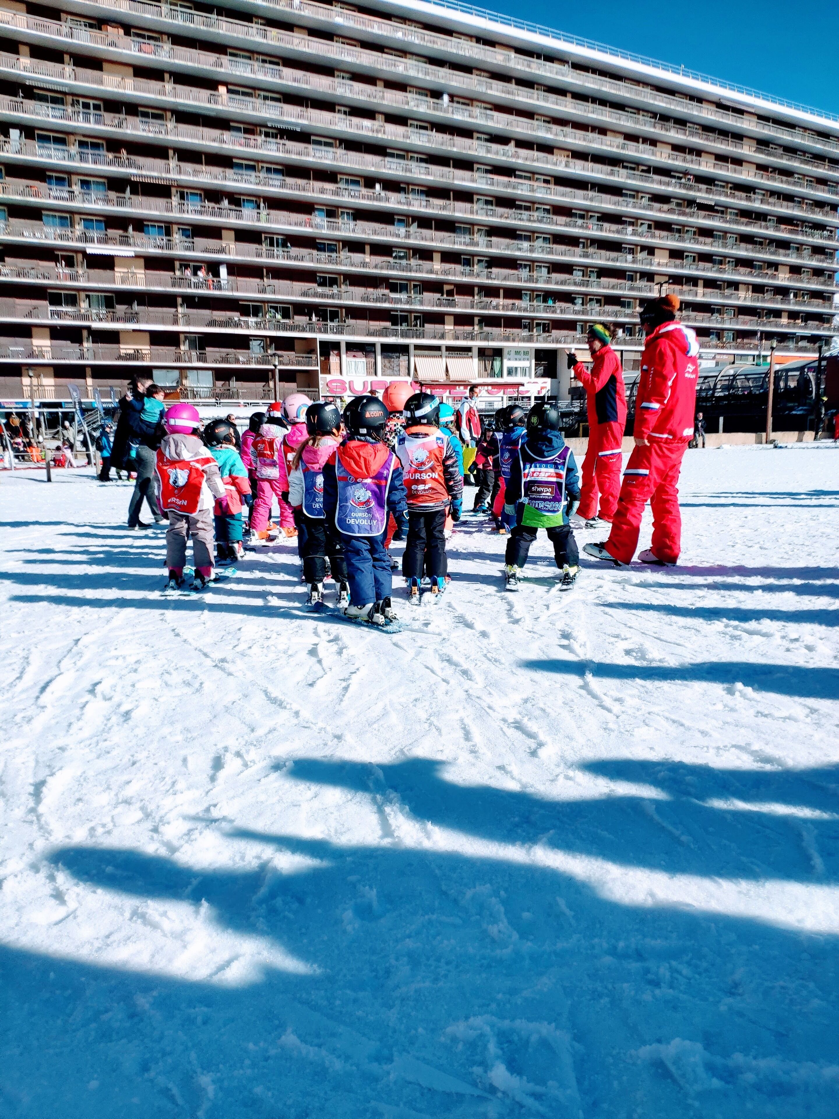 A group of children in winter sports attire gathered on a snowy surface with a large building in the background.