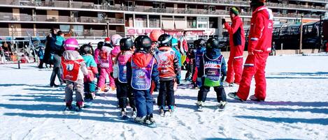 A group of children in winter sports attire gathered on a snowy surface with a large building in the background.