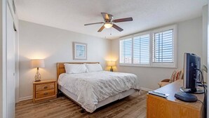 A bedroom with a bed, bedside lamps, a ceiling fan, a desk with a computer, and a window with blinds.