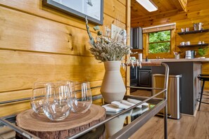Un intérieur en bois comprenant un coin cuisine, une table à manger en bois avec des verres et un vase, ainsi qu’une fenêtre donnant sur un paysage verdoyant.
