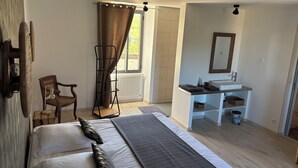 Une chambre avec un lit, une chaise, un étendoir, une fenêtre avec des rideaux, une salle de bain avec un lavabo et un miroir, et un radiateur.