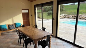 Un espace repas avec une table et des chaises en bois, un banc avec des coussins et une vue sur une piscine, visible à travers de grandes portes vitrées.