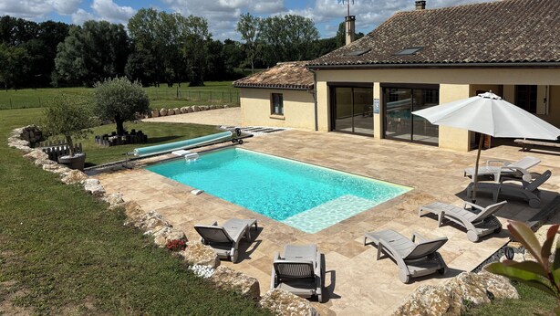 Une piscine dotée de chaises longues et d’un toboggan, entourée d’un jardin aménagé et d’une maison avec un toit en tuiles.