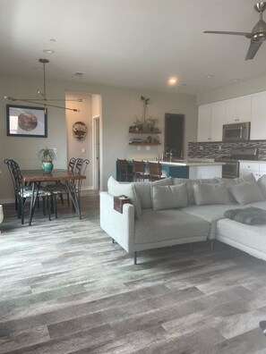 Un salon moderne avec un canapé d’angle, un coin repas comprenant des chaises et une table, une cuisine avec des meubles blancs et un four à micro-ondes, et un ventilateur de plafond.