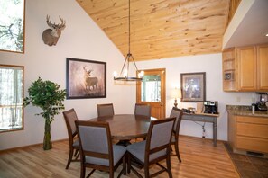 A dining area with a wooden table and chairs, a chandelier, and a mounted deer head on the wall. - The Duke Cabin - A FOREST LAKES Cabin in the Woods! HOT TUB (Coconino County)