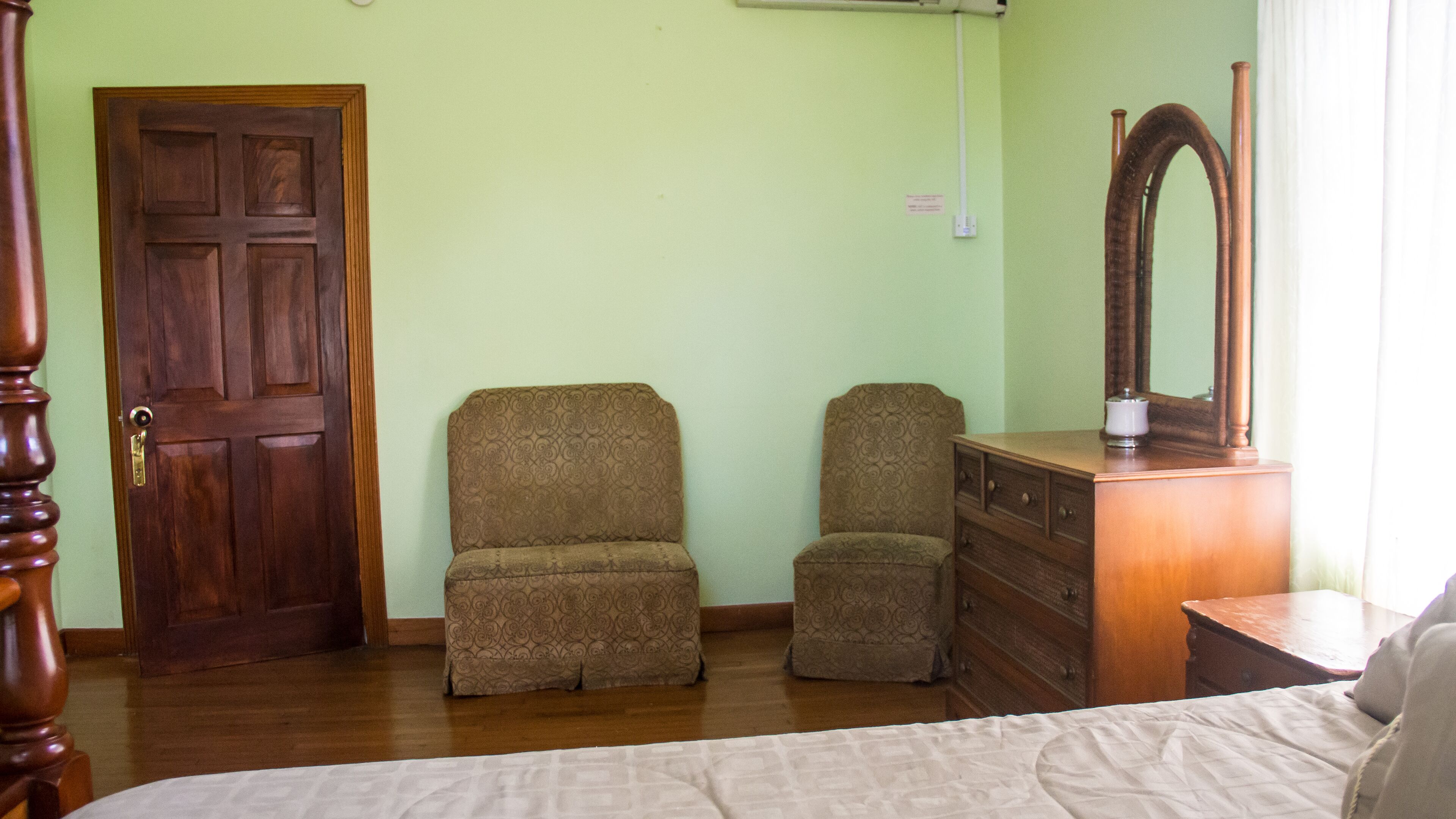 A bedroom with a wooden door, two patterned armchairs, a wooden dresser, and a bed with a white bedspread.